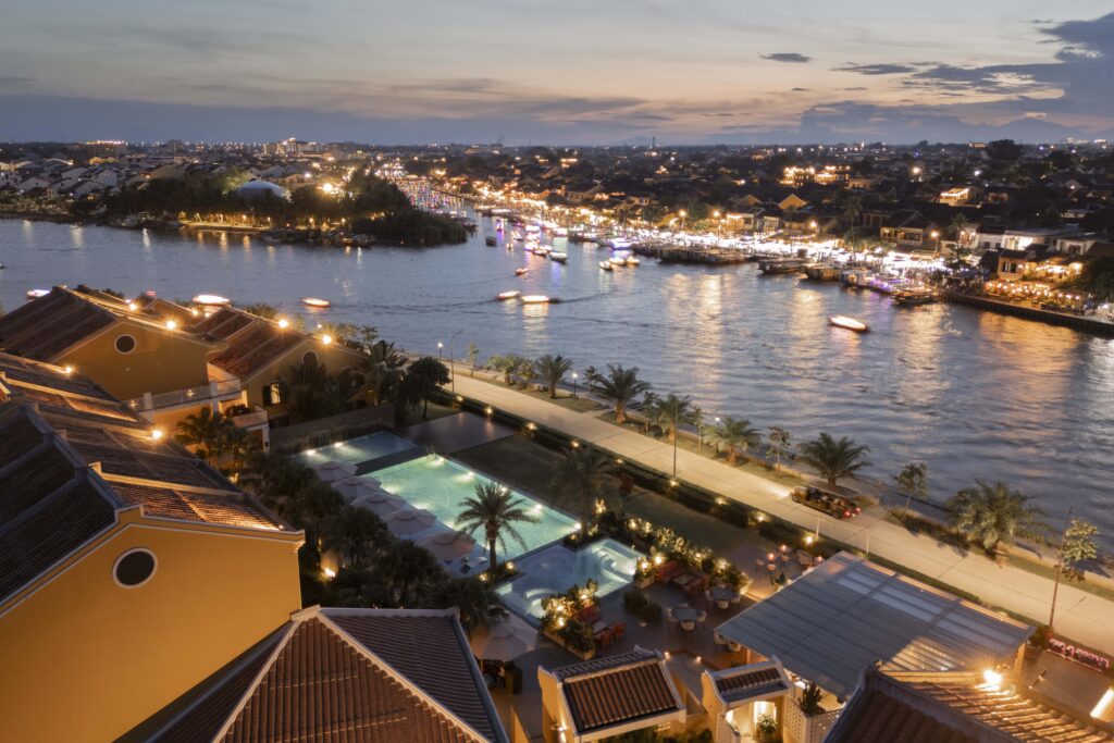 Aerial twilight view of Moire Hoi An, Vignette Collection by IHG, featuring its illuminated outdoor swimming pool and the Hoai River overlooking the lantern-lit Hoi An Ancient Town.